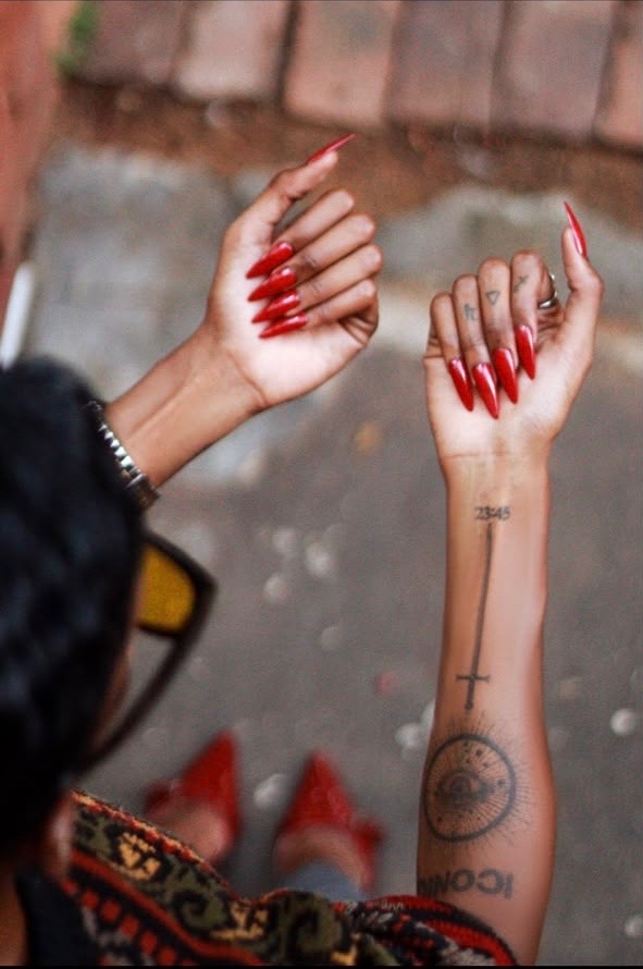 Red stiletto press-on nails, hands raised with wrist tattoos visible