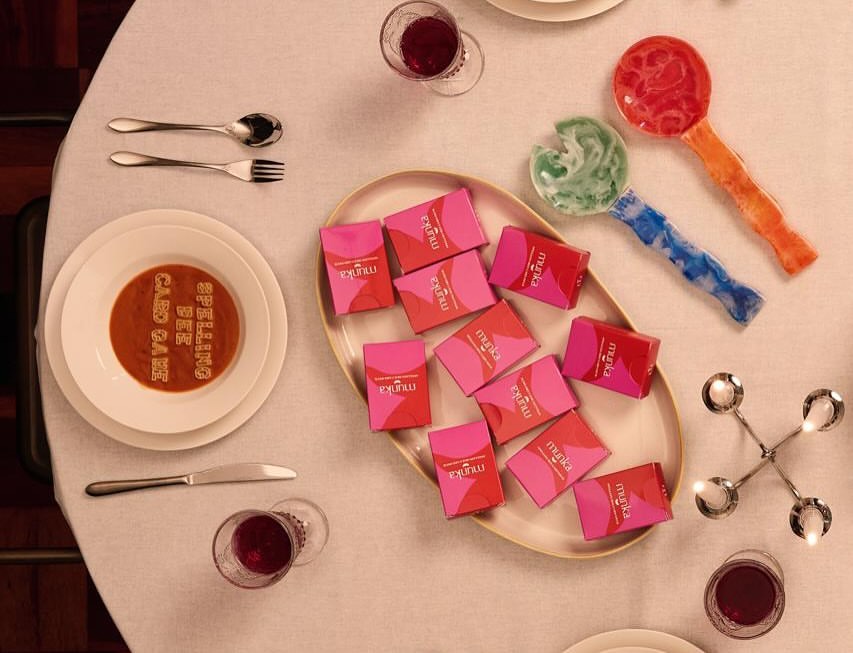 Overhead shot of Munka game boxes styled on a dinner table with wine glasses and a dessert reading 'Spelling Bee Card Game'
