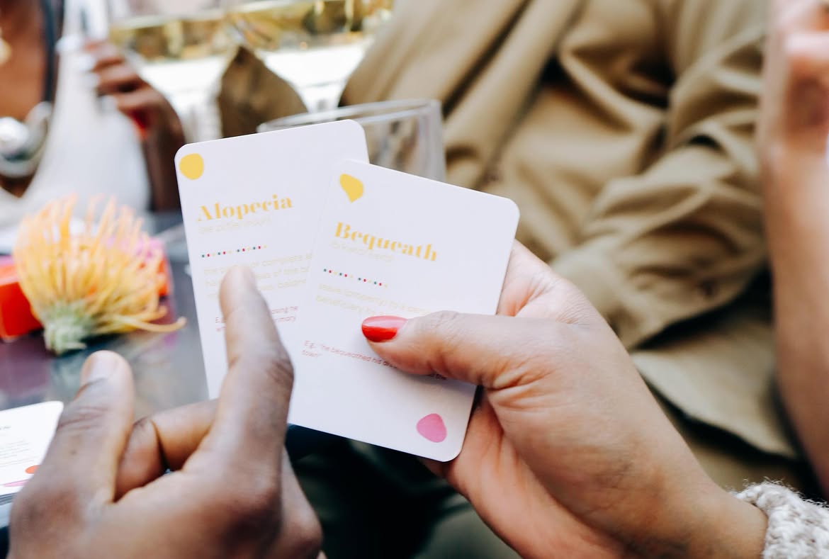 Close-up of hands holding Munka game cards with word definitions, 'Alopecia' and 'Bequeath'
